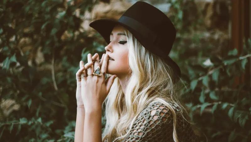 Subtle pagan jewelry on a blonde woman's hand. She is wearing a hat and surrounded by plants.