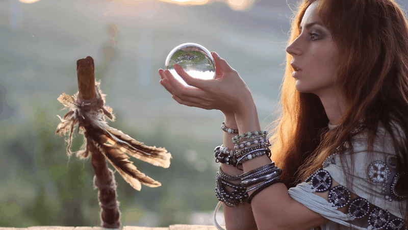 Decorative image of a psychic eclectic witch holding a crystal ball. In front of her is a staff with feathers on it.