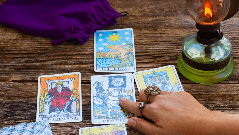 Decorative image of a woman pointing to the death tarot card in a tarot card spread on a wooden table next to a lamp and purple pouch