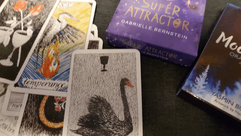 Decorative image of tarot cards and oracle cards. The tarot cards have a black swan and a heron. The oracle cards include the Super Attractor deck by Gabrielle Bernstein and the Moonology deck.
