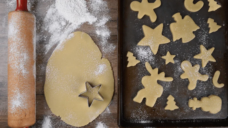 A rolling pin dusted with flour. Sugar cookie dough rolled out with a star cookie cutter on it. Sugar cookies shaped like reindeer, gingerbread men, angels, stars, trees, and snowmen.