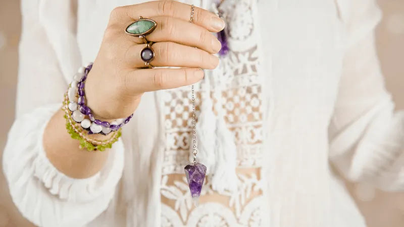 A woman with an amethyst pendulum wearing a preppy sweater and many crystal bracelets and rings