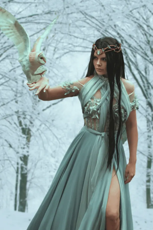 A sorceress in the forest with her trusty owl familiar wearing a sheer white dress and a diadem. Her long black hair trails down her sides.