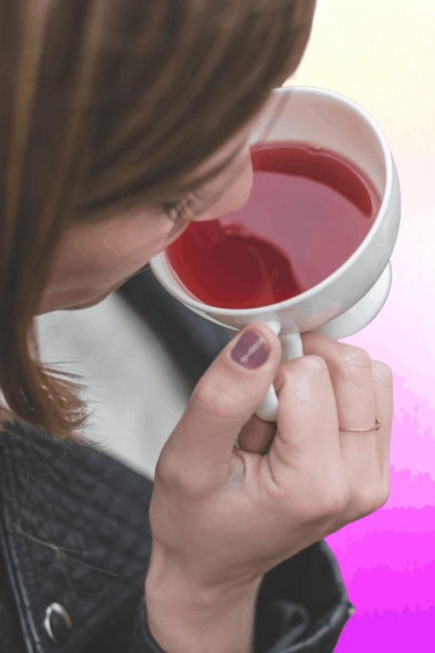A woman drinking mugwort tea on a white and pink background. Her nail is purple and she is wearing a thin minimalist ring,