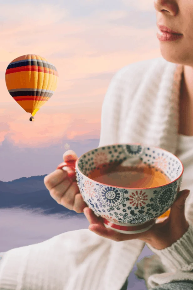 A woman drinking mugwort tea in a white robe while look out at a dreamlike landscape with a hot air balloon