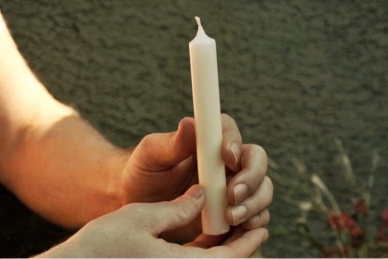 Two hands cradling white taper candle vertically, soft green foliage background, showing pre-blessing hand position