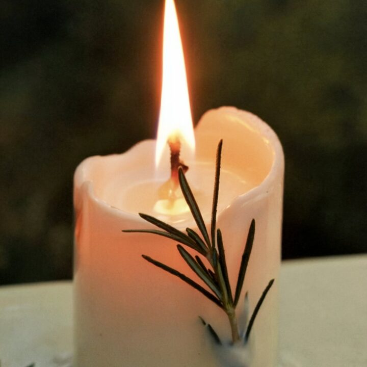 White pillar candle burning with fresh rosemary sprig decoration, bright flame casting glow, showing blessed candle use