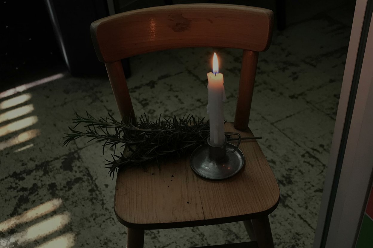 White candle and fresh rosemary on a wooden kitchen chair show how witchcraft happens in regular homes with ordinary objects. The empty chair signals presence through absence, a common pattern in folk magic where what's missing matters as much as what's there. Kitchen witchery uses whatever's available: grocery store herbs, dollar store candles, furniture as altar.