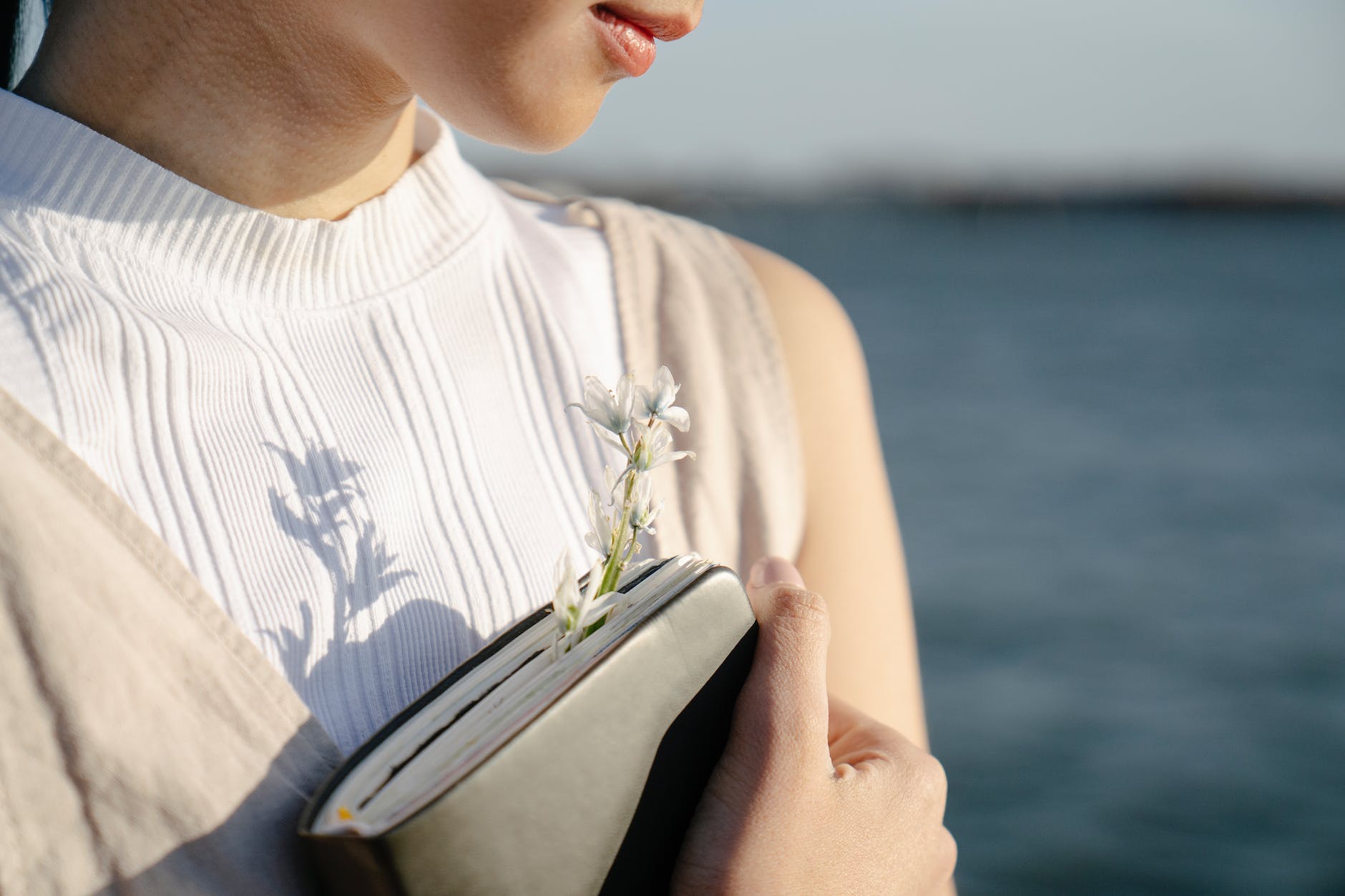 crop romantic woman with blooming flower in notebook against lake