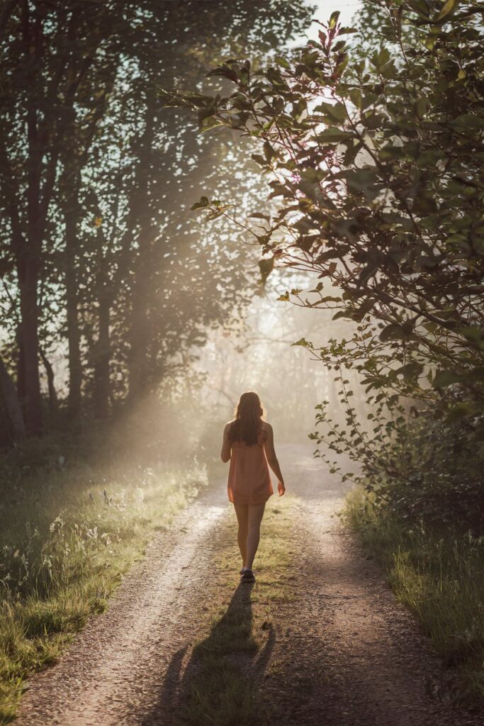 Abstract depiction of a person walking through a peaceful forest path, with sunlight filtering through trees, representing her need for space and natural reflection. Visual cue: earthy tones, feminine, soul-nurturing vibe