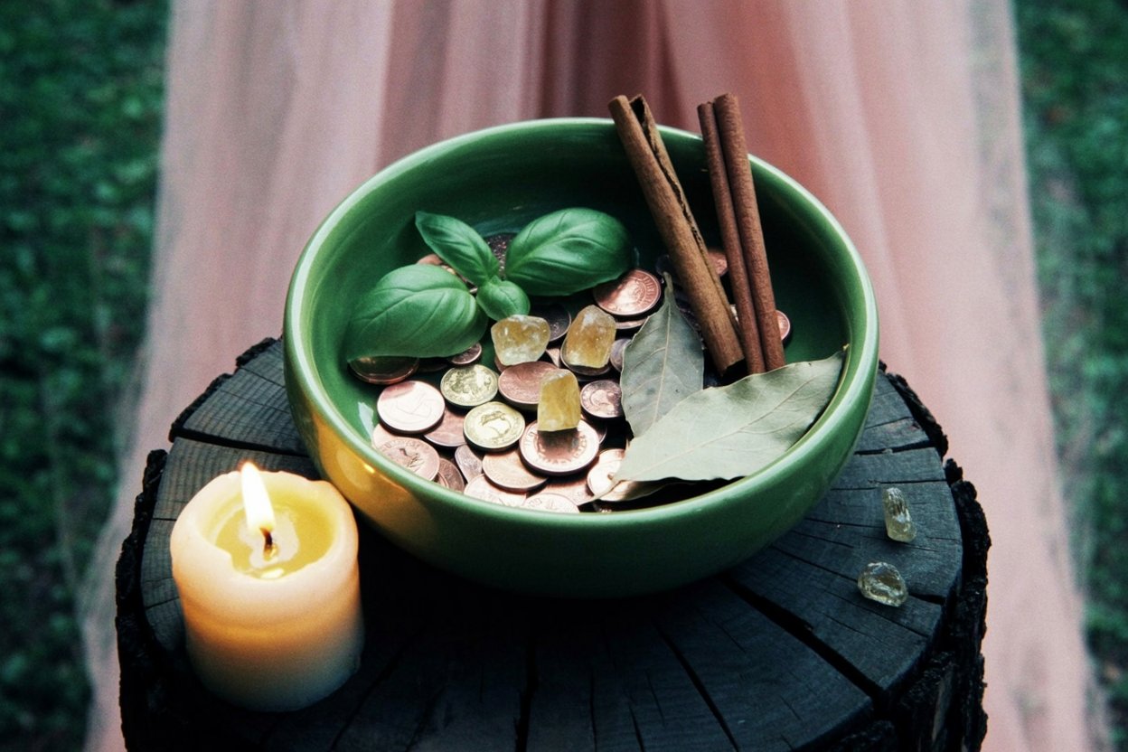 Money bowl photographed during active ritual work with lit candle providing both illumination and ongoing energy feeding. The combination of coins in copper and silver tones, fresh green basil, dried bay leaves, and cinnamon sticks shows the layering of multiple prosperity correspondences working together. The natural wood stump base grounds the magical work in earth element stability.