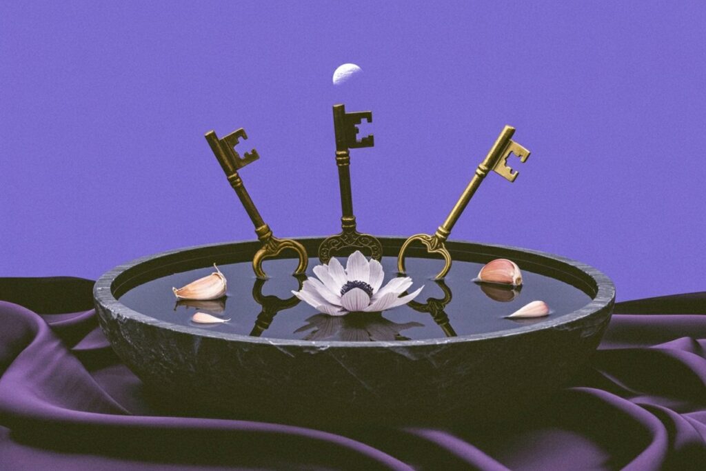 Three ornate brass and bronze skeleton keys rest in a dark ceremonial bowl with floating white anemone and garlic cloves beneath a crescent moon, representing Hekate's role as keeper of keys that unlock hidden knowledge and her connection to lunar cycles and protective magic.
