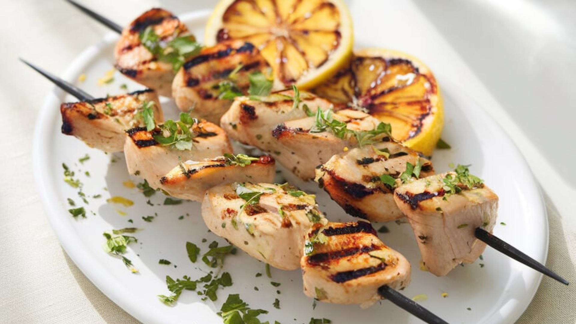 Grilled lemon herb chicken skewers served on a pristine white plate, with beautifully charred lemon slices at the side. The skewers feature juicy, golden-brown chicken pieces sprinkled with chopped fresh parsley. The natural sunlight highlights the dish’s textures, creating a vibrant and inviting atmosphere perfect for a Lughnasadh feast.