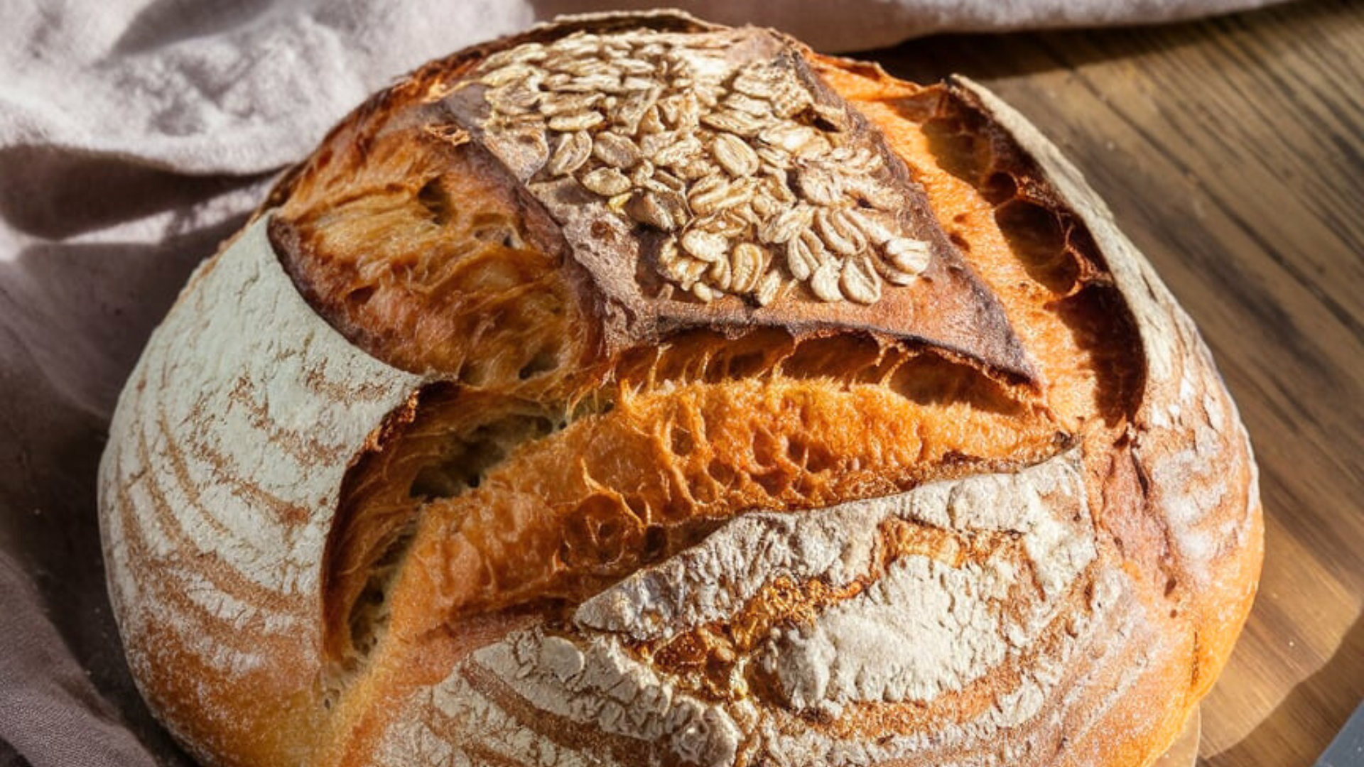 Homemade bread cutting board details A close-up of a perfectly baked honey oat sourdough loaf resting on a rustic wooden cutting board. The crust is golden and slightly cracked, with a generous topping of toasted oats. A soft beige linen napkin is casually draped beside it, with a sharp wooden-handled bread knife resting nearby. The setting is bathed in warm, natural light that highlights the bread's texture and rich color.