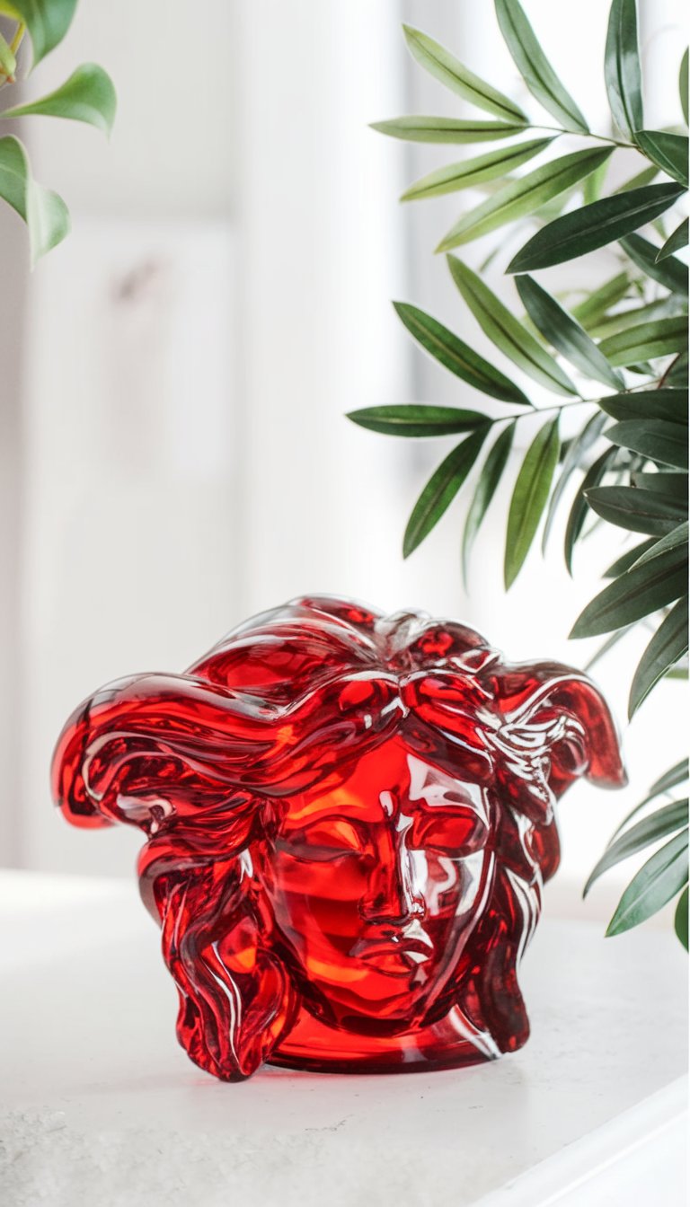 Glossy red crystal Versace Medusa paperweight displayed on a white surface, surrounded by lush green leaves. The intricate details of the iconic Medusa head catch the light, creating a striking blend of luxury, boldness, and elegance in a modern decor setting.