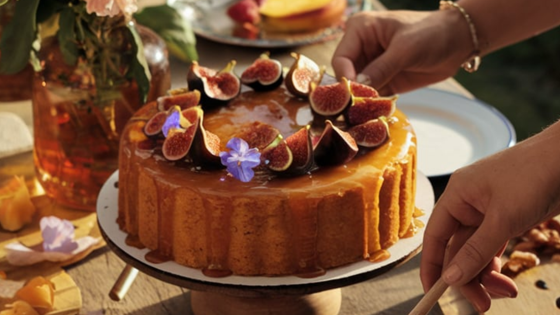 A golden honey-glazed cake sits atop a wooden cake stand, adorned with fresh figs and delicate edible flowers. Warm sunlight highlights the rich caramel drizzle as hands reach in to serve a slice.