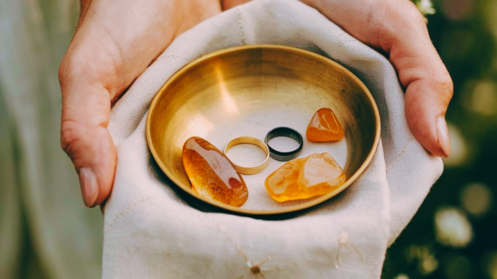 This image shows a close-up of a person holding a small brass bowl on a soft, off-white cloth. Inside the bowl are two rings—one gold and one black—along with several polished amber-colored stones. The scene is set outdoors with blurred greenery in the background, giving it a natural and intimate feel. The warm tones of the brass, rings, and stones complement the earthy and romantic aesthetic, evoking a timeless and symbolic moment, possibly related to a wedding or ritual.