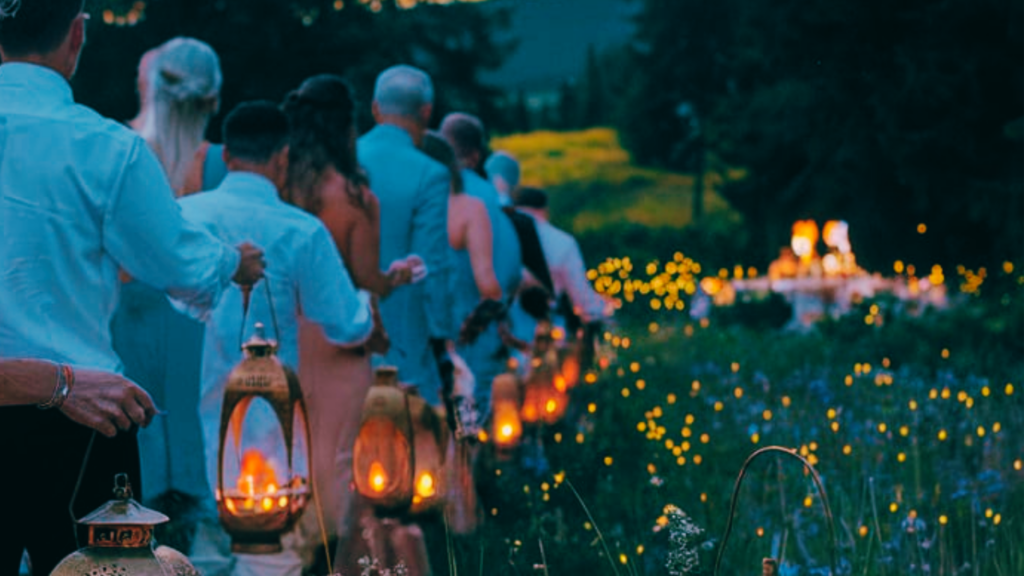 A line of wedding guests in elegant attire moves through a twilight meadow, each carrying an ornate brass lantern. The soft glow of candlelight and the distant glow of a wedding reception in the background create a fairytale-like atmosphere.
