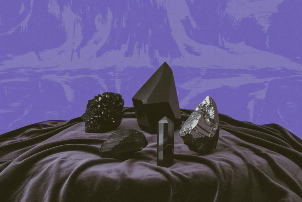 A collection of black crystals arranged on deep purple velvet fabric with dramatic folds. From left: a cubic black tourmaline cluster with matte rough surface, a tall hexagonal black tourmaline prism showing vertical striations, a metallic silver-gray hematite specimen with reflective finish, and several smaller rough black crystal pieces. This arrangement shows a working Hecate altar stone collection. Tourmaline establishes protective boundaries, hematite provides grounding after underworld journeying, and varied crystal forms serve different threshold work. You accumulate these over time as you learn what you actually need, not all at once. She teaches you to reach for the right tool when the moment asks for it.