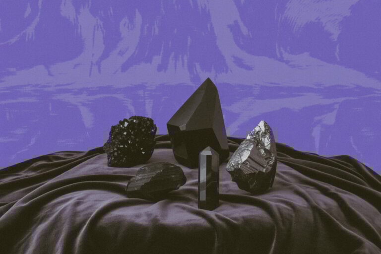 A collection of black crystals arranged on deep purple velvet fabric with dramatic folds. From left: a cubic black tourmaline cluster with matte rough surface, a tall hexagonal black tourmaline prism showing vertical striations, a metallic silver-gray hematite specimen with reflective finish, and several smaller rough black crystal pieces. This arrangement shows a working Hecate altar stone collection. Tourmaline establishes protective boundaries, hematite provides grounding after underworld journeying, and varied crystal forms serve different threshold work. You accumulate these over time as you learn what you actually need, not all at once. She teaches you to reach for the right tool when the moment asks for it.