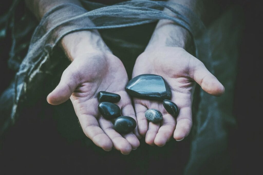 Hands holding six black obsidian stones: large angular piece right palm, smaller rounded stones left. Weight teaches grounding after underworld journeys.