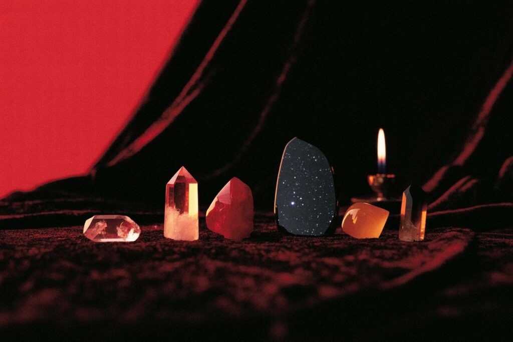 Six crystals arranged left to right on burgundy velvet with candle flame. Shows Venus dawn stones and Mars fire stones used together on working altar.