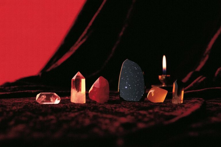 Six crystals arranged left to right on burgundy velvet with candle flame. Shows Venus dawn stones and Mars fire stones used together on working altar.